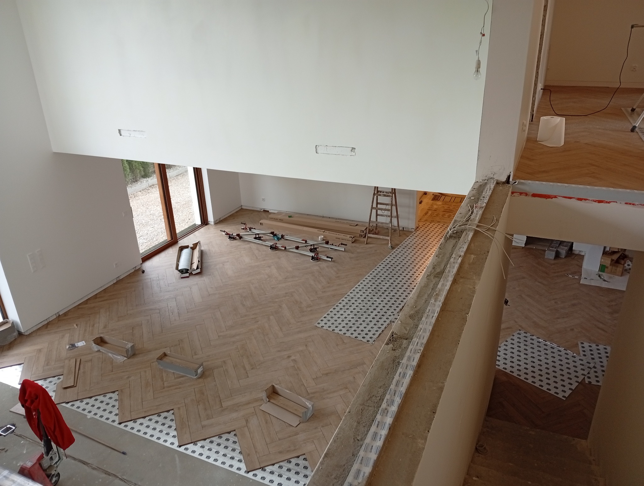 Układanie parkietu w jodełkę w przestronnym, jasnym wnętrzu. Widoczne narzędzia, klej i częściowo ułożona podłoga. Ujęcie z góry, z widokiem na okno i schody.