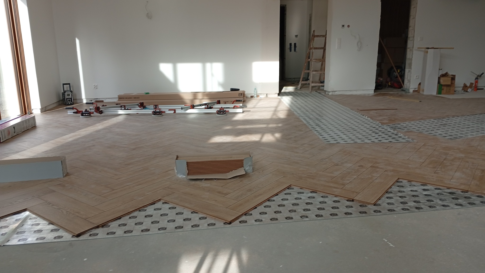 Układanie parkietu w jodełkę. Widoczne panele, podkład i narzędzia. Częściowo ułożona podłoga w jasnym pomieszczeniu z dużym oknem i drabiną w tle.