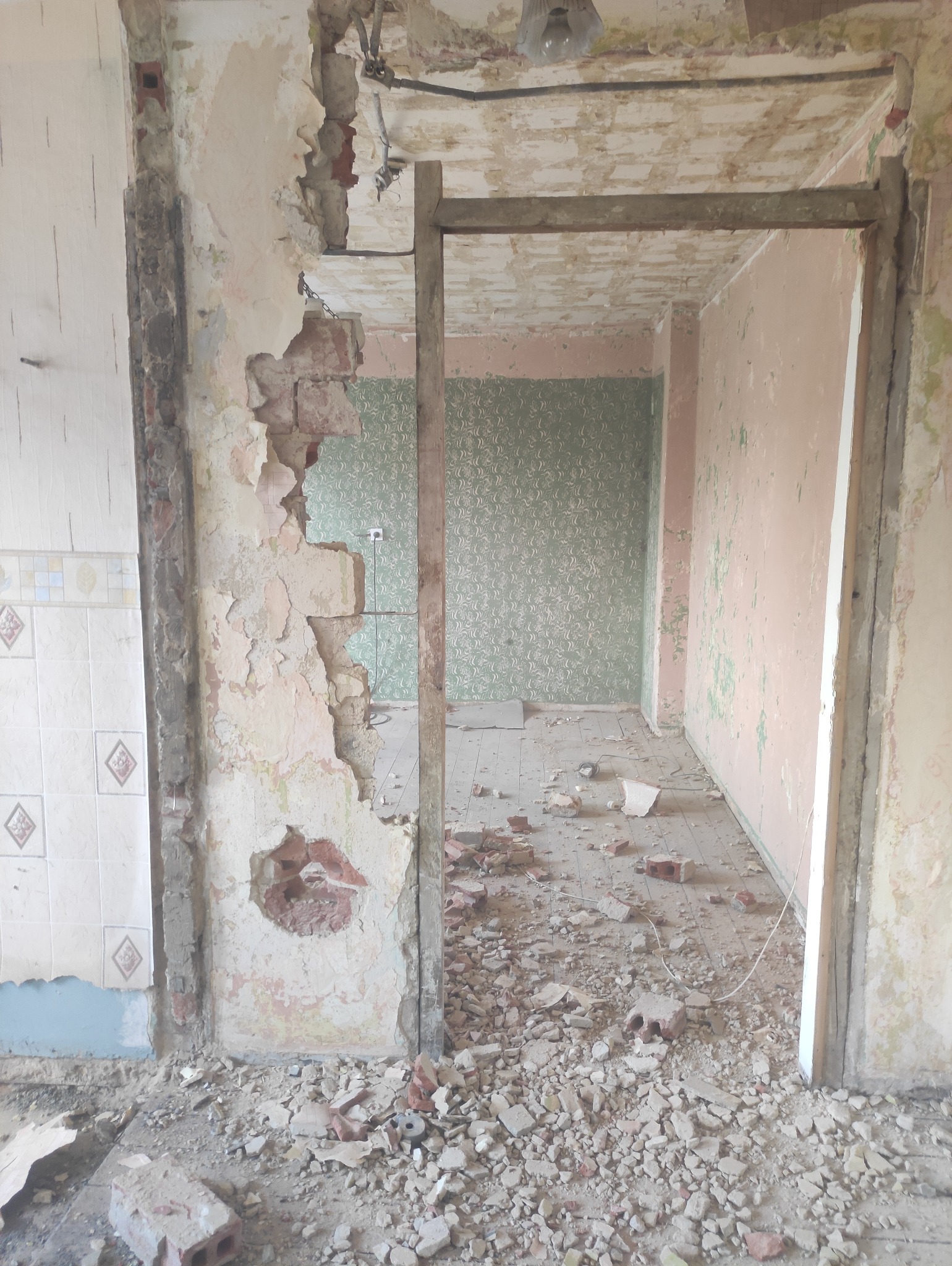 Wnętrze w trakcie rozbiórki: fragment ściany z odsłoniętą cegłą, gruz na podłodze, widoczne stare tapety i elementy instalacji. Ujęcie z bliska, surowy stan.
