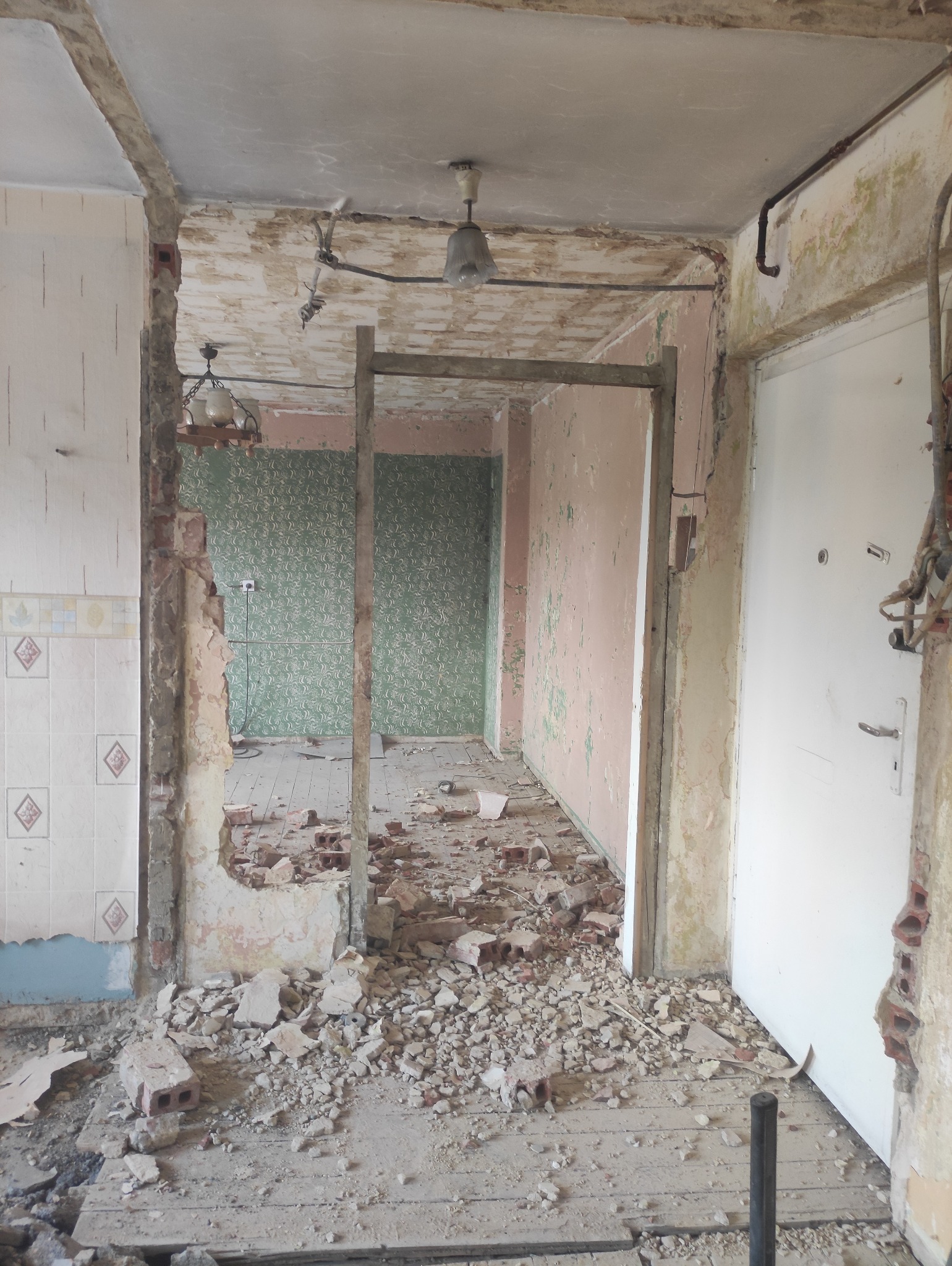 Wnętrze mieszkania w trakcie rozbiórki: gruz na podłodze, odsłonięte ściany z fragmentami tapet i płytek, widoczne instalacje. Ujęcie z perspektywy wejścia.