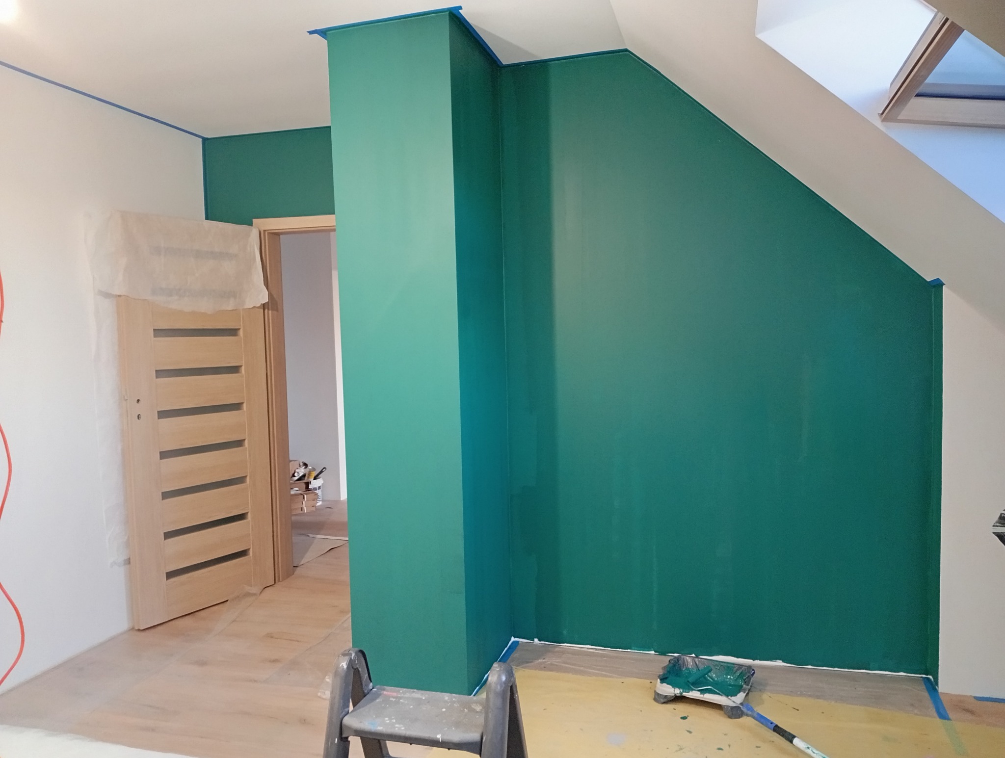 Ściana pomalowana na zielono w pokoju ze skosami, zabezpieczona taśmą malarską. Widoczna drabina malarska i kuweta z farbą. Drzwi oklejone folią ochronną.
