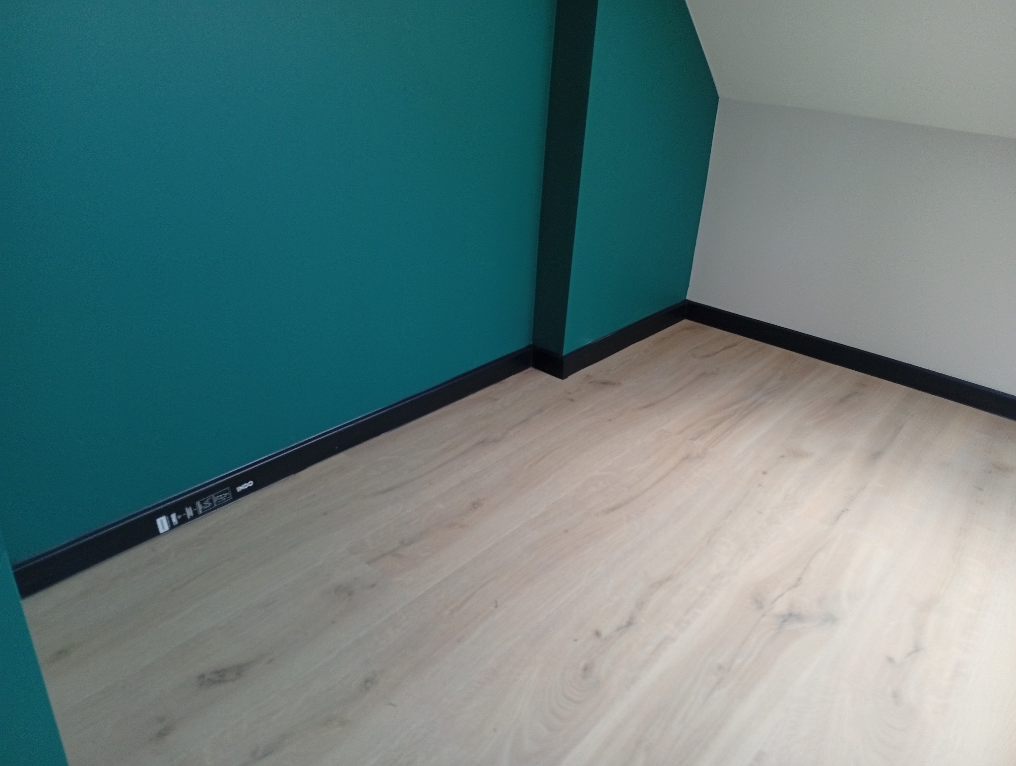 Ściana pomalowana na turkusowo, kontrastująca z białą ścianą i jasną podłogą z paneli. Czarne listwy przypodłogowe dodają elegancji. Widoczny fragment kolumny.