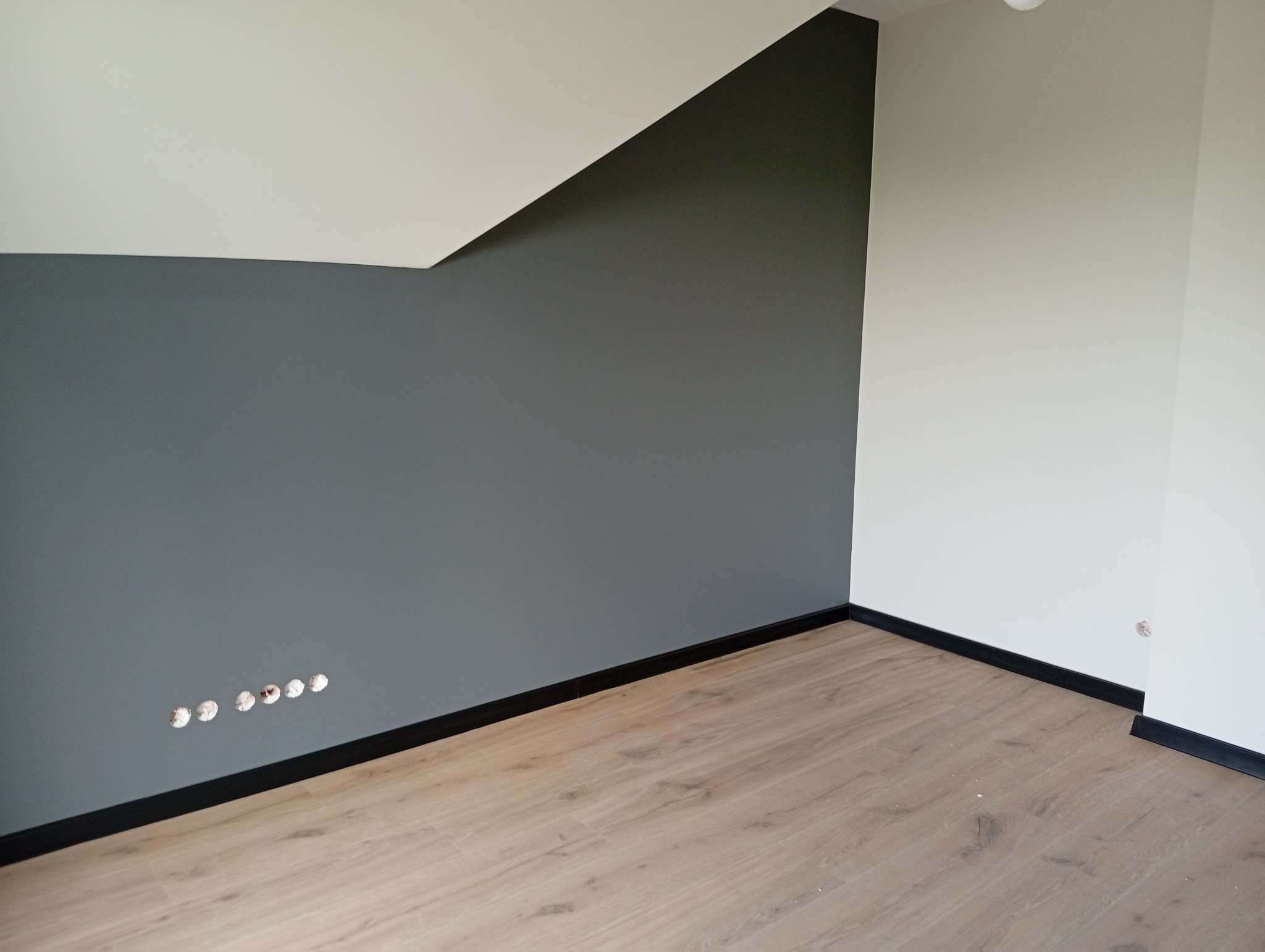 Wnętrze po malowaniu: szara ściana z gniazdkami, biała ściana, drewniana podłoga i czarne listwy przypodłogowe. Ukośny sufit dodaje charakteru.