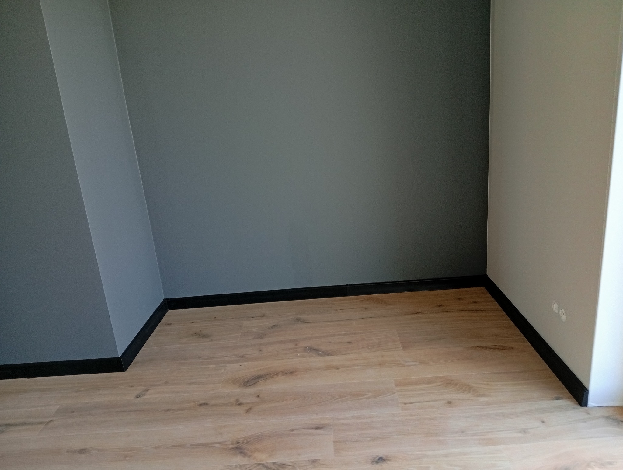 Wnętrze po remoncie: świeżo ułożone panele podłogowe w odcieniach drewna, kontrastujące z szarą ścianą i czarnymi listwami przypodłogowymi. Widoczne gniazdka elektryczne.
