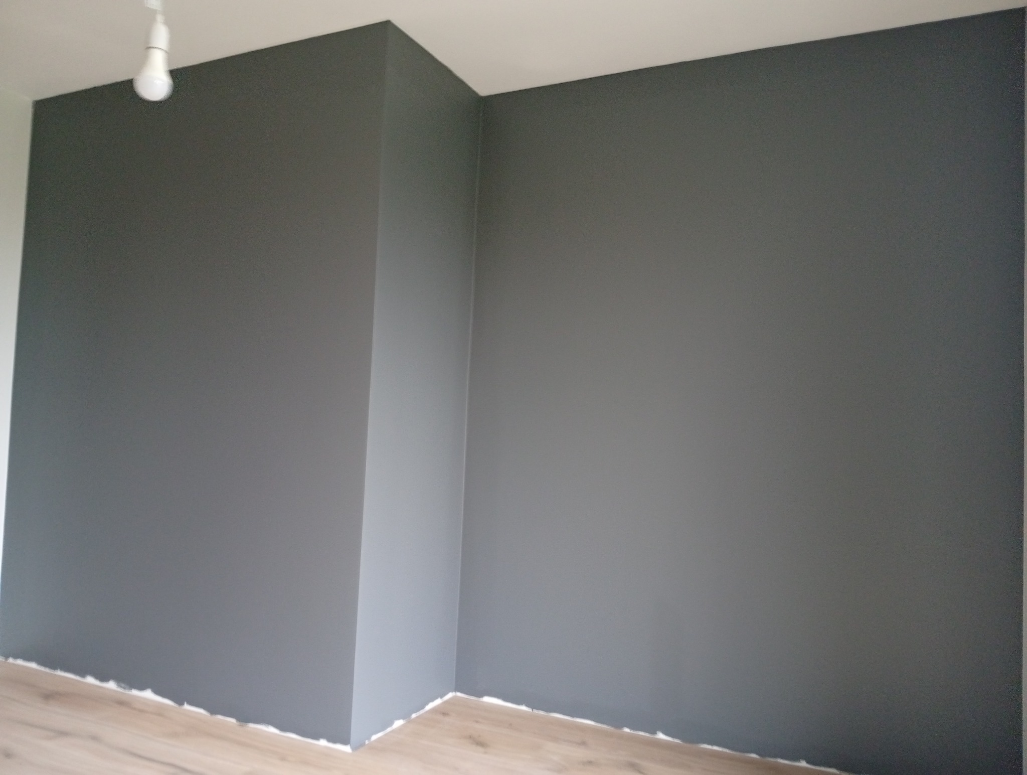 Świeżo pomalowany pokój w odcieniach szarości, z widoczną żarówką i fragmentem drewnianej podłogi. Minimalistyczne wnętrze po remoncie.