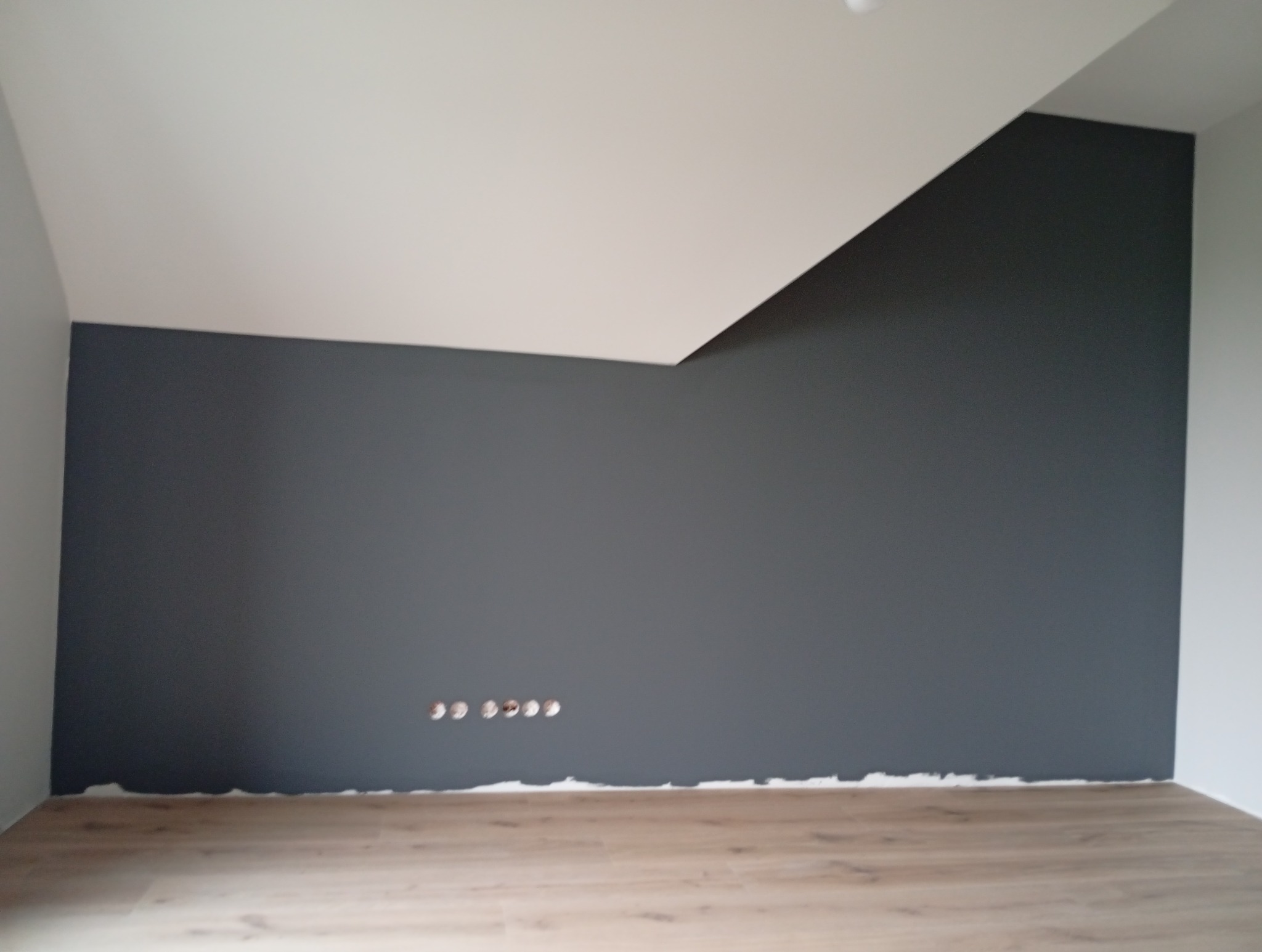 Ściana pomalowana na szaro z widocznymi otworami na gniazdka elektryczne i fragmentem podłogi z paneli. Biały sufit z nietypowym załamaniem.