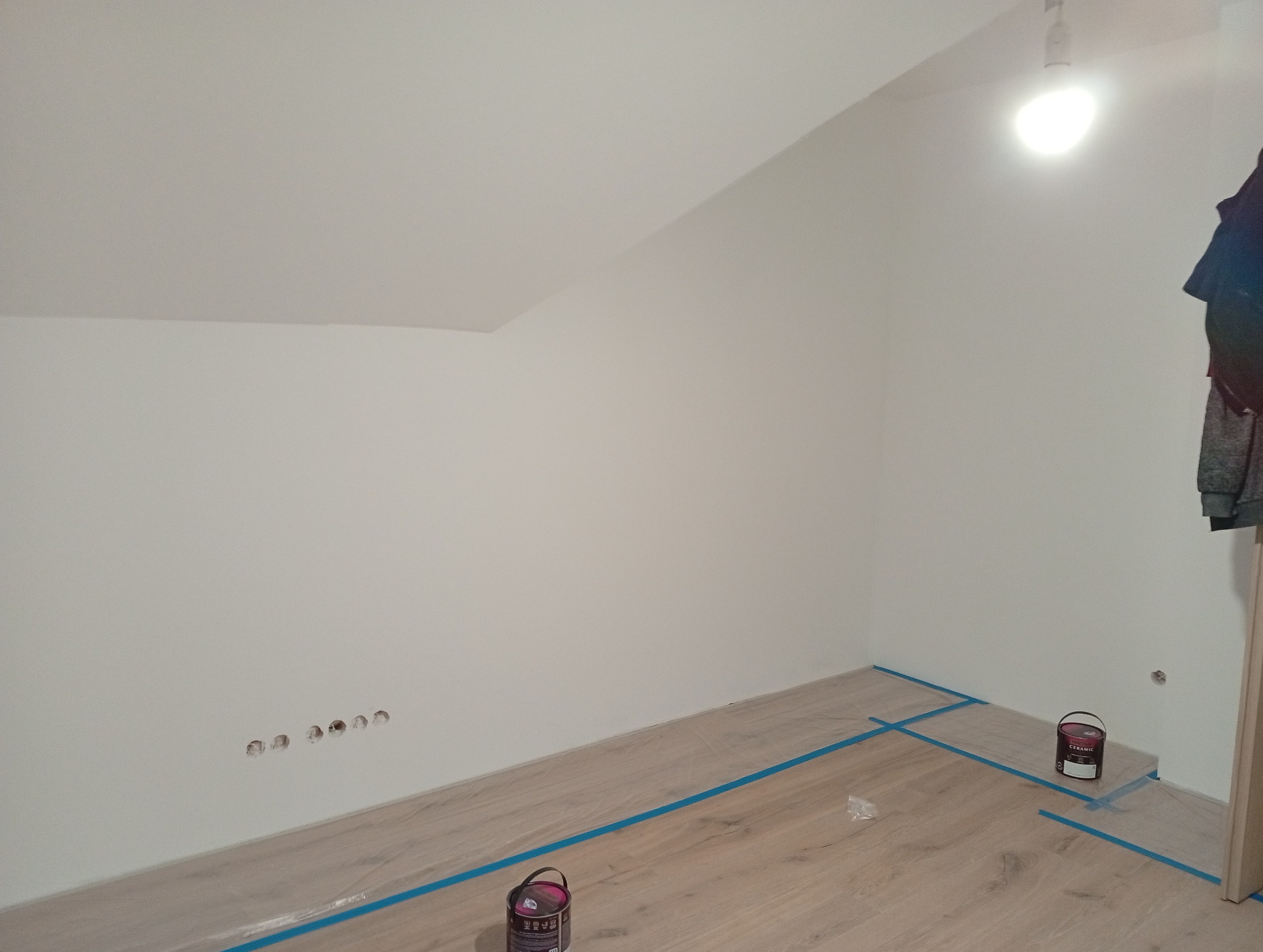 Przygotowane do malowania poddasze w Zabrzu: zabezpieczona podłoga panelowa, puszki z farbą ceramiczną, gładkie, białe ściany i sufit ze skosem.