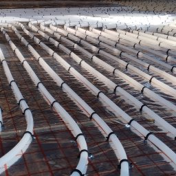 BULER THERMAL ENERGY Piotr Buler - Układ ogrzewania podłogowego z białych rur mocowanych czarnymi uchwytami na folii z siatką, widok z perspektywy, oświetlenie naturalne.