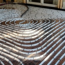 BULER THERMAL ENERGY Piotr Buler - Białe rury ogrzewania podłogowego ułożone spiralnie na folii izolacyjnej, przymocowane czarnymi klipsami do siatki. Widoczny fragment ściany i okna.