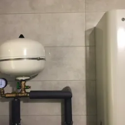 Biały zbiornik wyrównawczy ciśnienia z manometrem, podłączony do rur z izolacją, obok białego bojlera LG na tle szarych płytek ściennych.