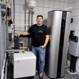 BULER THERMAL ENERGY Piotr Buler - Instalacja gruntowej pompy ciepła M-TEC WPS26 w kotłowni z białymi kafelkami. Mężczyzna w czarnym T-shircie stoi obok pompy. Widoczne rury i zbiornik wyrównawczy.