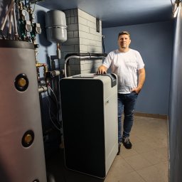 BULER THERMAL ENERGY Piotr Buler - Instalacja gruntowej pompy ciepła M-TEC WPD412 w kotłowni. Widoczny zbiornik buforowy, orurowanie i instalator w białej koszulce. Ściany wyłożone płytkami.