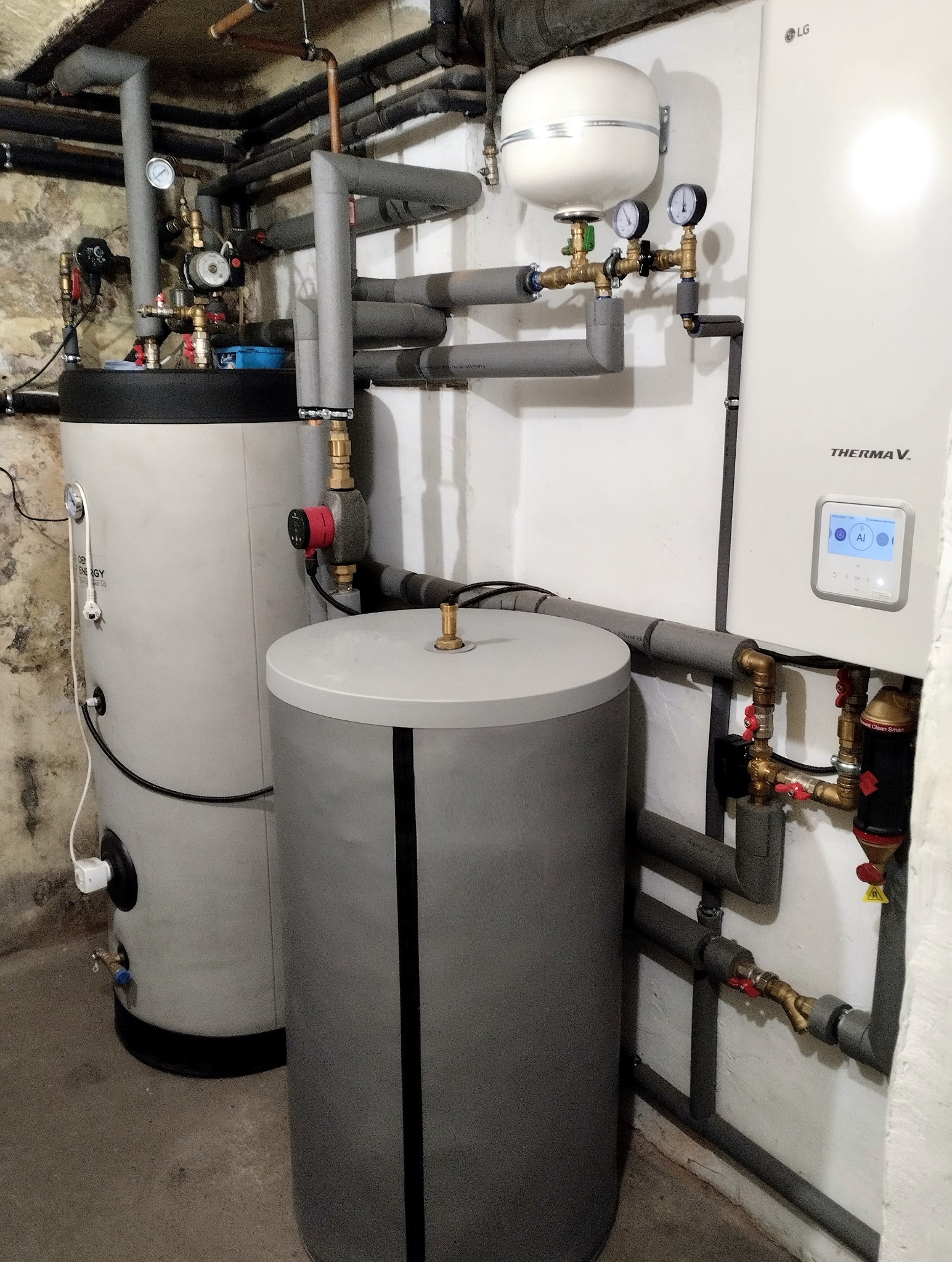 Pompa ciepła LG Therma V w kotłowni z grzejnikami. Widoczne zbiorniki i rury z izolacją, manometry. Ściany częściowo z surowego betonu.