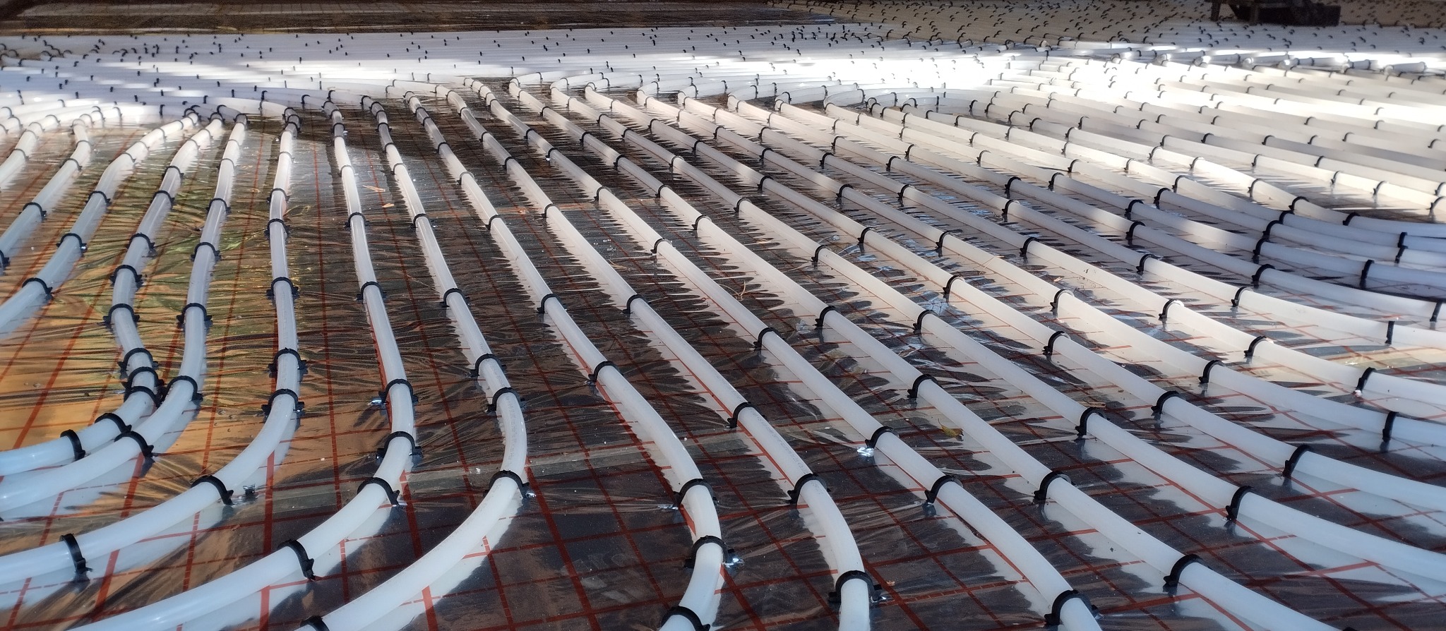 Układ ogrzewania podłogowego z białych rur mocowanych czarnymi uchwytami na folii z siatką, widok z perspektywy, oświetlenie naturalne.