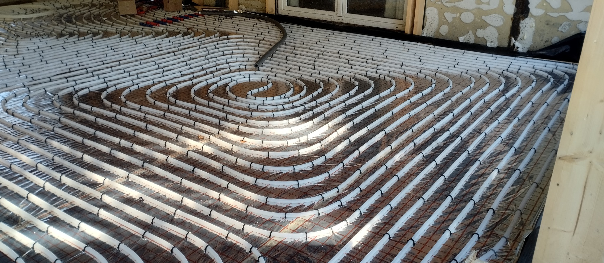 Białe rury ogrzewania podłogowego ułożone spiralnie na folii izolacyjnej, przymocowane czarnymi klipsami do siatki. Widoczny fragment ściany i okna.