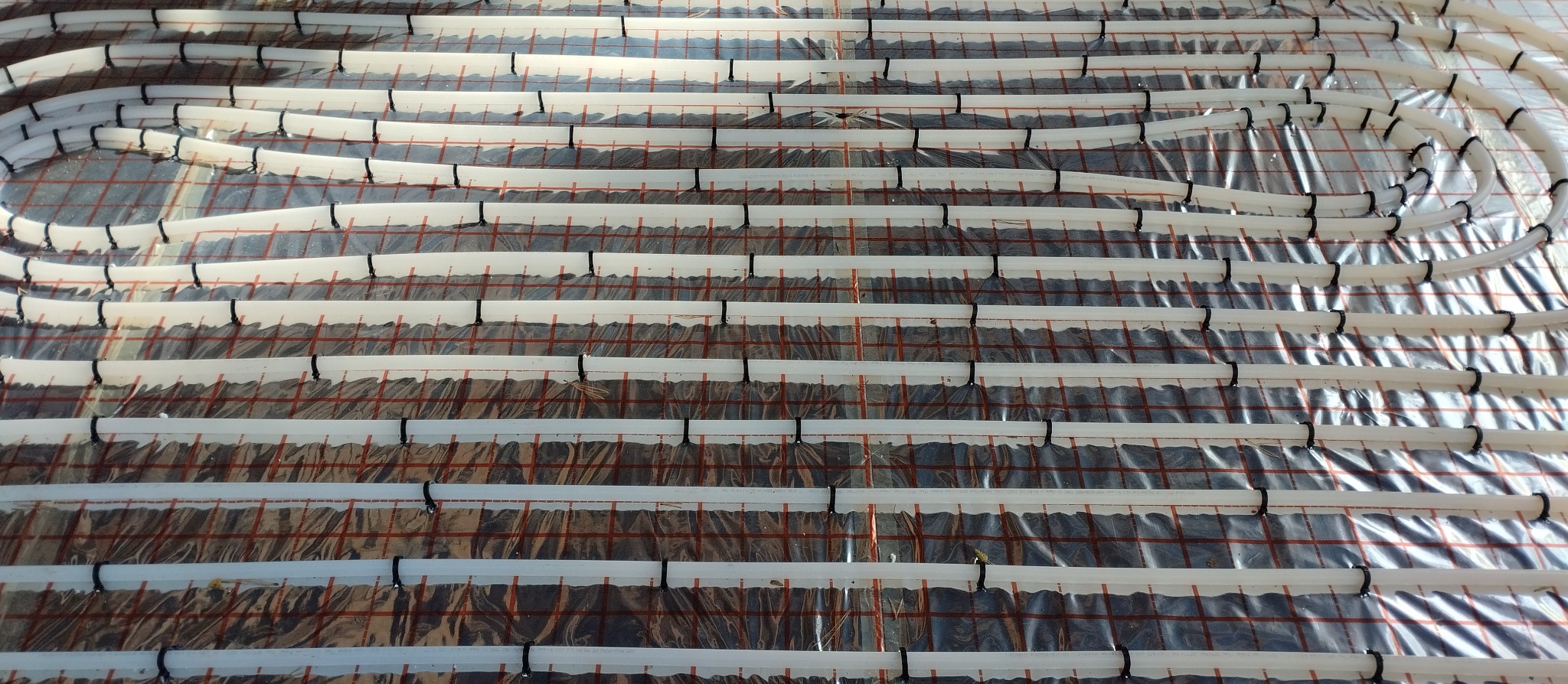 Białe rury ogrzewania podłogowego, równolegle ułożone na folii izolacyjnej z czerwoną siatką, przymocowane czarnymi uchwytami. Widok z góry.