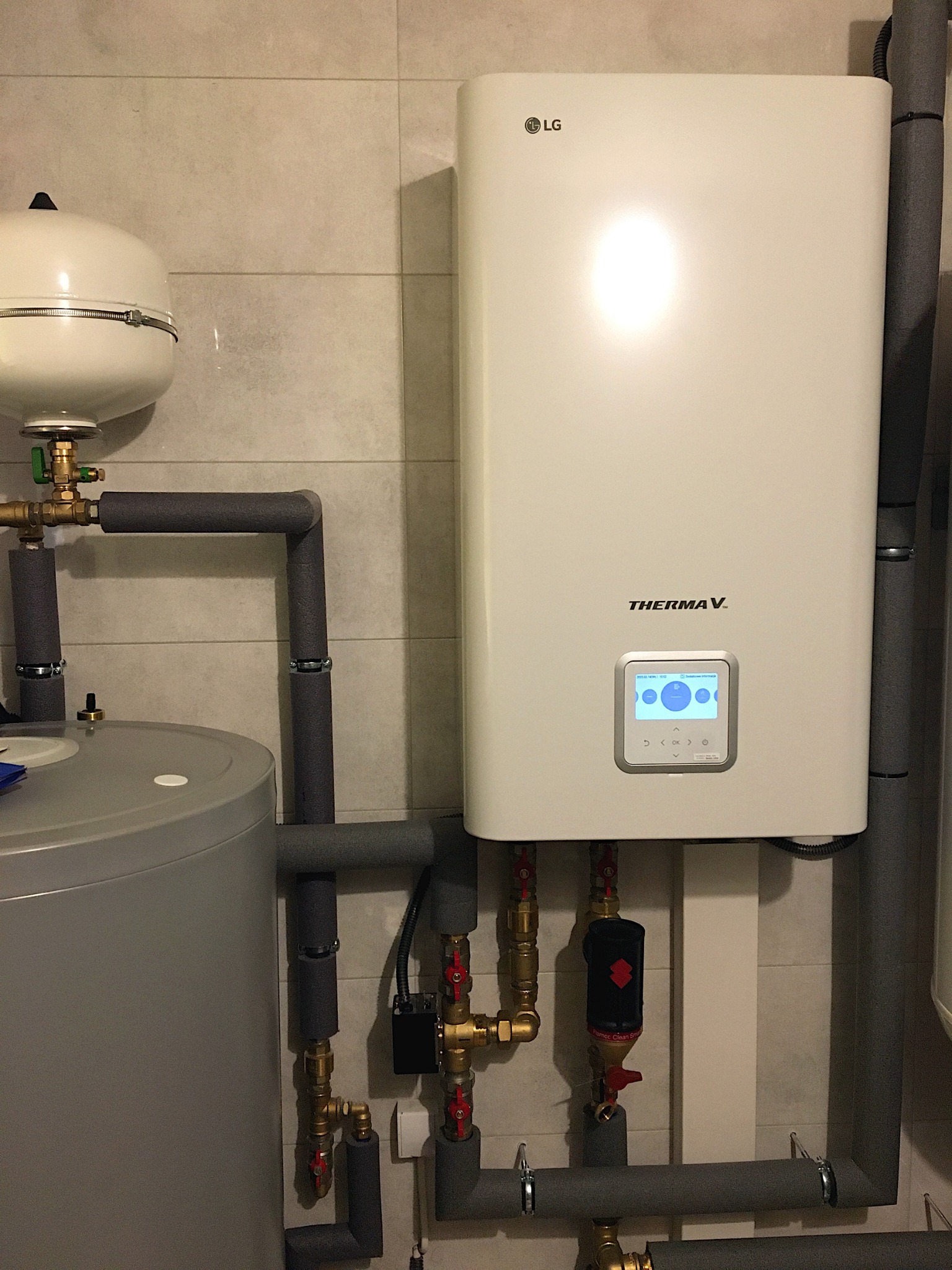 Biała pompa ciepła LG Therma V z szarymi rurami PEX i mosiężną armaturą na tle jasnych płytek. Widoczny zbiornik C.W.U. i separator Flamco.