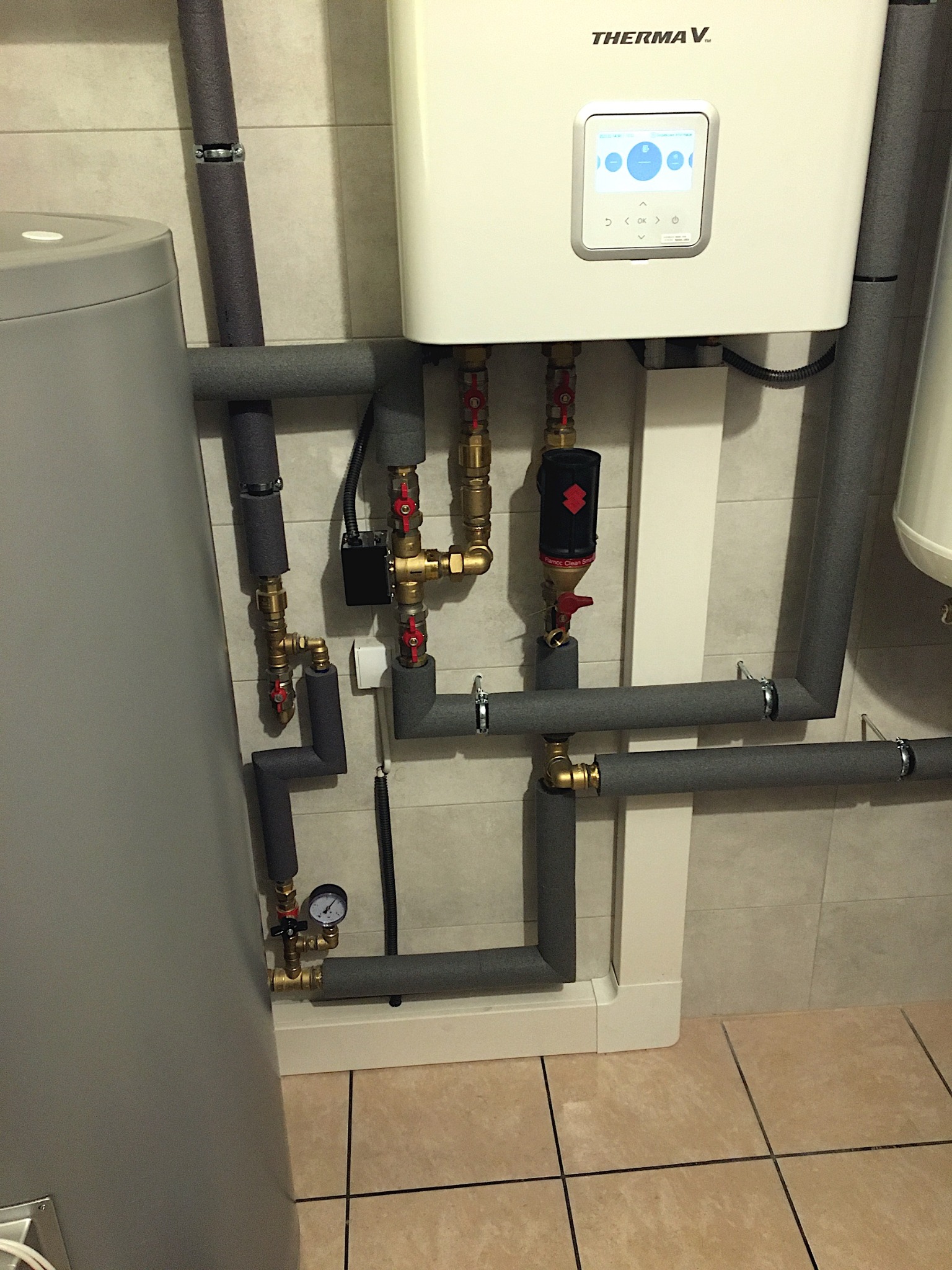 Biały piec grzewczy Therma V z szarymi, zaizolowanymi rurami i armaturą na tle beżowych płytek. Widoczny zbiornik i manometr.