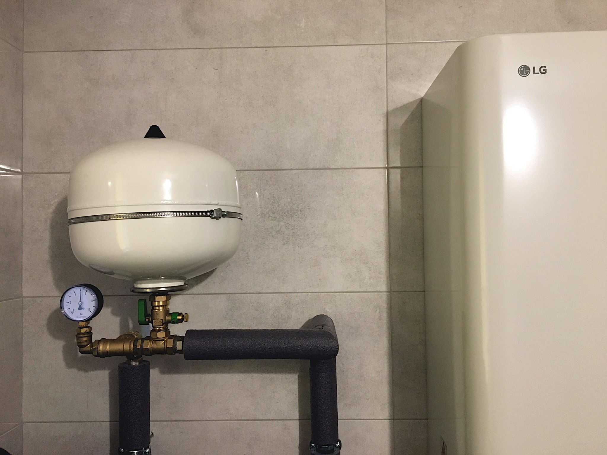Biały zbiornik wyrównawczy ciśnienia z manometrem, podłączony do rur z izolacją, obok białego bojlera LG na tle szarych płytek ściennych.