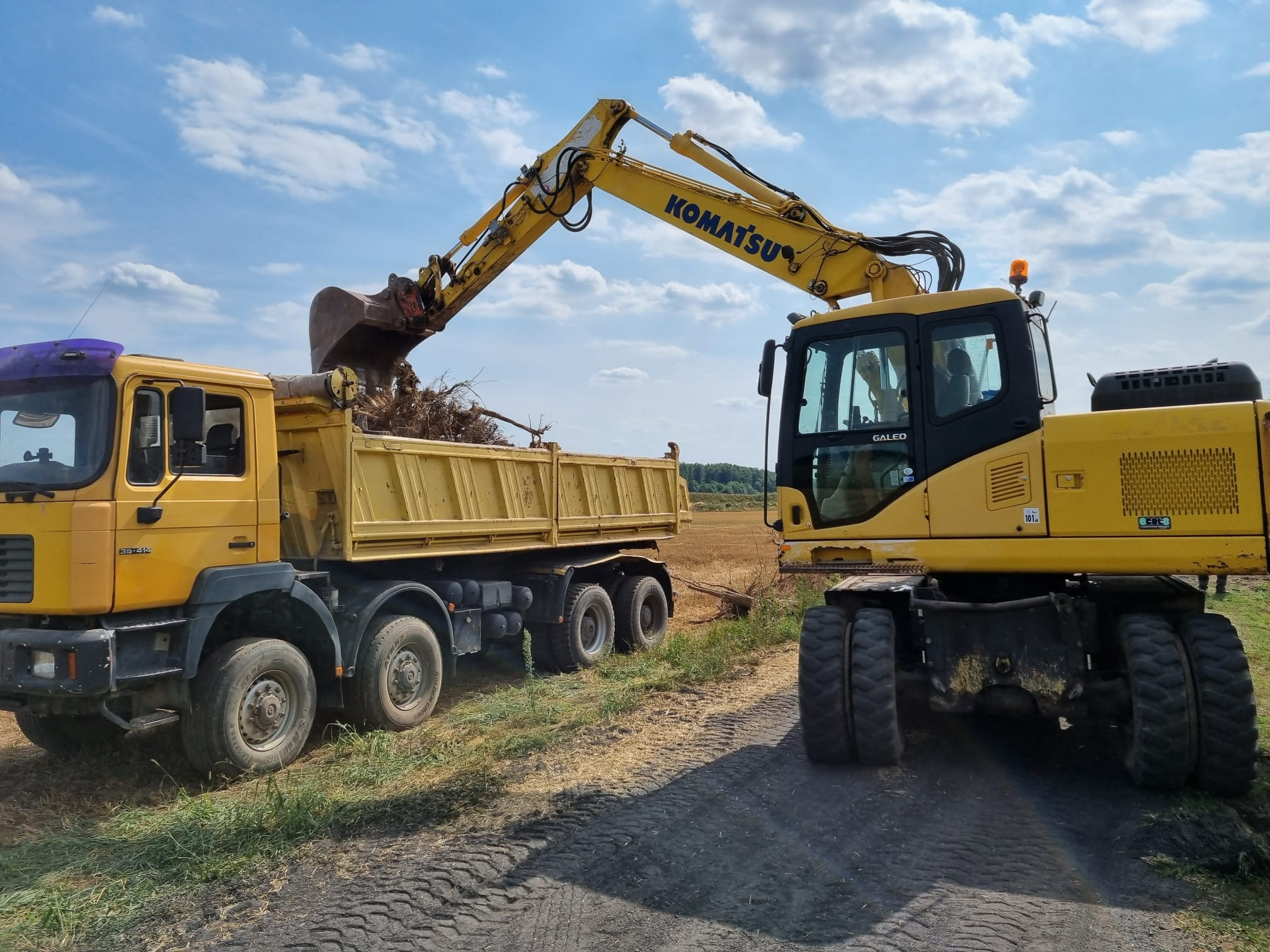 Żółta koparka Komatsu załadowuje korzenie i gałęzie do żółtej ciężarówki MAN na tle błękitnego nieba z chmurami.