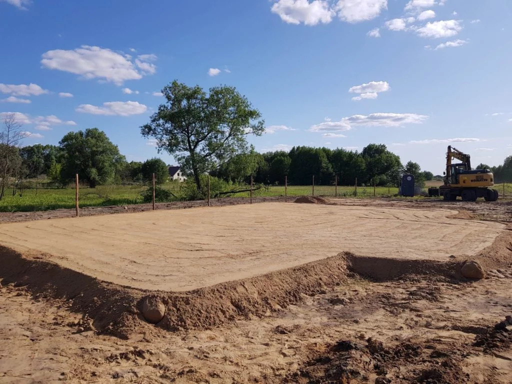 Przygotowany teren pod budowę z wytyczonym obrysem fundamentów, widoczny żółty sprzęt budowlany i niebieskie niebo z chmurami.