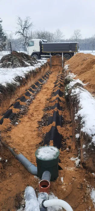 Długi wykop w ziemi zabezpieczony czarną geowłókniną, z widoczną studzienką kontrolną i rurami drenażowymi; w tle ciężarówka wywożąca urobek na zaśnieżonym terenie.