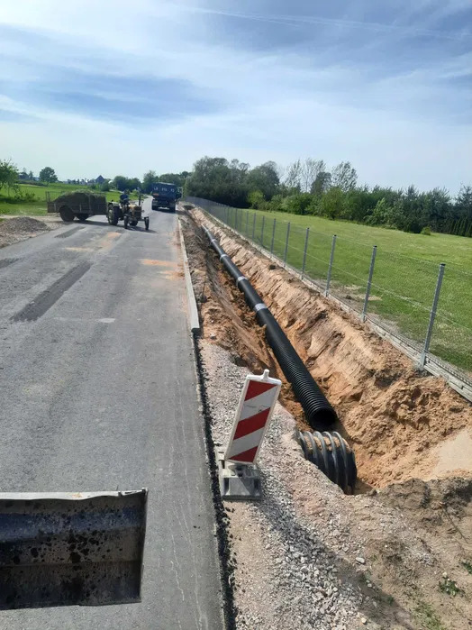 Montaż czarnej rury drenażowej w wykopie wzdłuż asfaltowej drogi, z widocznym traktorem z przyczepą i ciężarówką w tle.