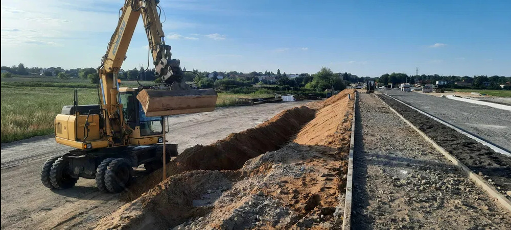 Żółta koparka kołowa z łyżką nabiera ziemię z wykopu wzdłuż nowej drogi z podsypką, widok na krajobraz z zabudowaniami w tle.