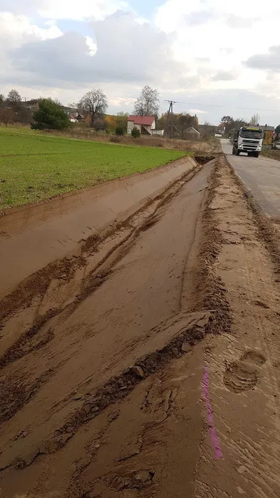 Świeżo wykopany rów melioracyjny wzdłuż drogi, z widocznymi śladami opon i fragmentem różowej farby na ziemi, na tle zielonego pola i zabudowań wiejskich, z ciężarówką w oddali.