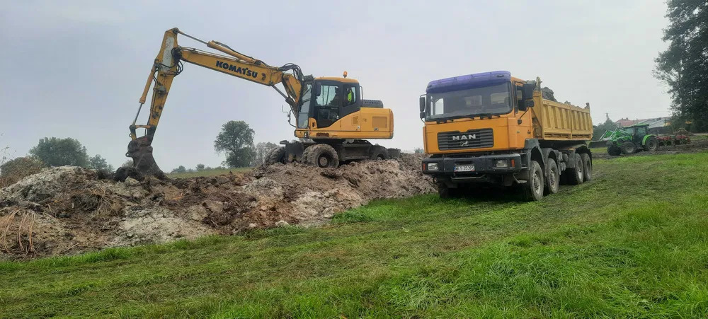 Żółta koparka Komatsu na grząskim terenie, obok wywrotka MAN, w tle zielony traktor. Prace ziemne w toku, pochmurne niebo.