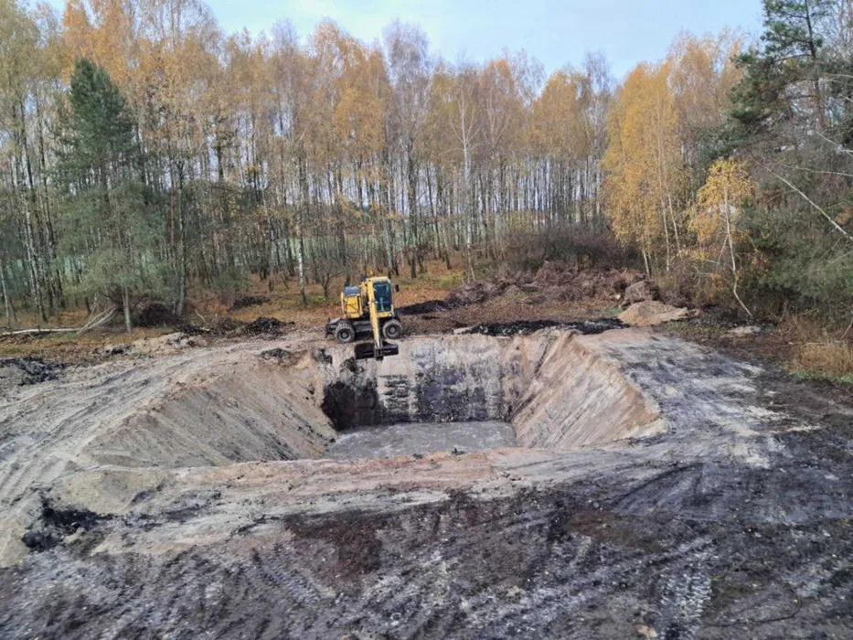 Żółta koparka na skraju wykopu w kształcie kwadratu wypełnionego wodą, otoczonego ziemią i drzewami w jesiennej scenerii.