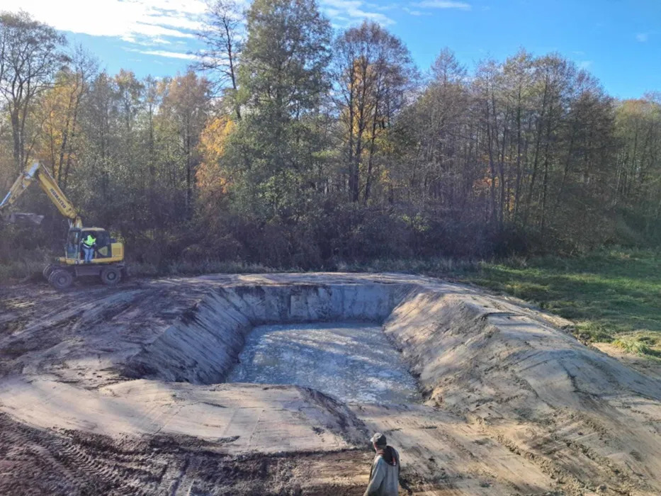 Wykopany, prostokątny dół wypełniony wodą, otoczony usypaną ziemią, z widoczną koparką Komatsu w tle i pracownikiem w dolnym rogu, teren podmokły, jesienne drzewa w tle.