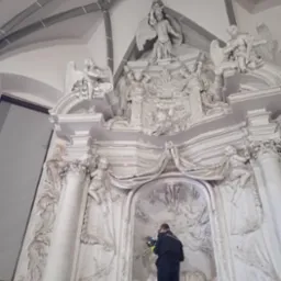 Pracownik w czarnym kombinezonie, w masce, czyści delikatne, białe rzeźby sakralne w kościele, używając narzędzia z niebieską końcówką, na rusztowaniu.