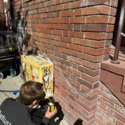 Pracownik w czarnej bluzie RadiusLabs z bliska czyści ceglaną elewację budynku przy użyciu specjalistycznego urządzenia, obok stoi żółta skrzynka z naklejkami i butelka wody.