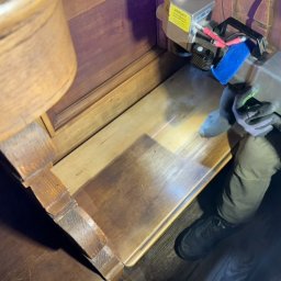 RADIUS LABS SP. Z O.O. | CZYSZCZENIE LASEREM - Laserowe czyszczenie fragmentu drewnianej szafki zabytkowej, widoczny sprzęt specjalistyczny w rękach pracownika, obszar poddawany obróbce wyraźnie jaśniejszy.