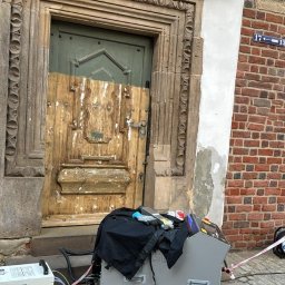 RADIUS LABS SP. Z O.O. | CZYSZCZENIE LASEREM - Stara drewniana brama z kamiennym obramowaniem w trakcie renowacji, z widocznym sprzętem do czyszczenia i zabezpieczającą taśmą.