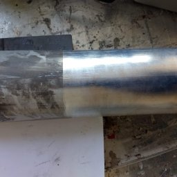 RADIUS LABS SP. Z O.O. | CZYSZCZENIE LASEREM - Metalowy element cylindryczny, w połowie oczyszczony z zabrudzeń, leżący na białym arkuszu papieru na tle betonowej podłogi z plamami.