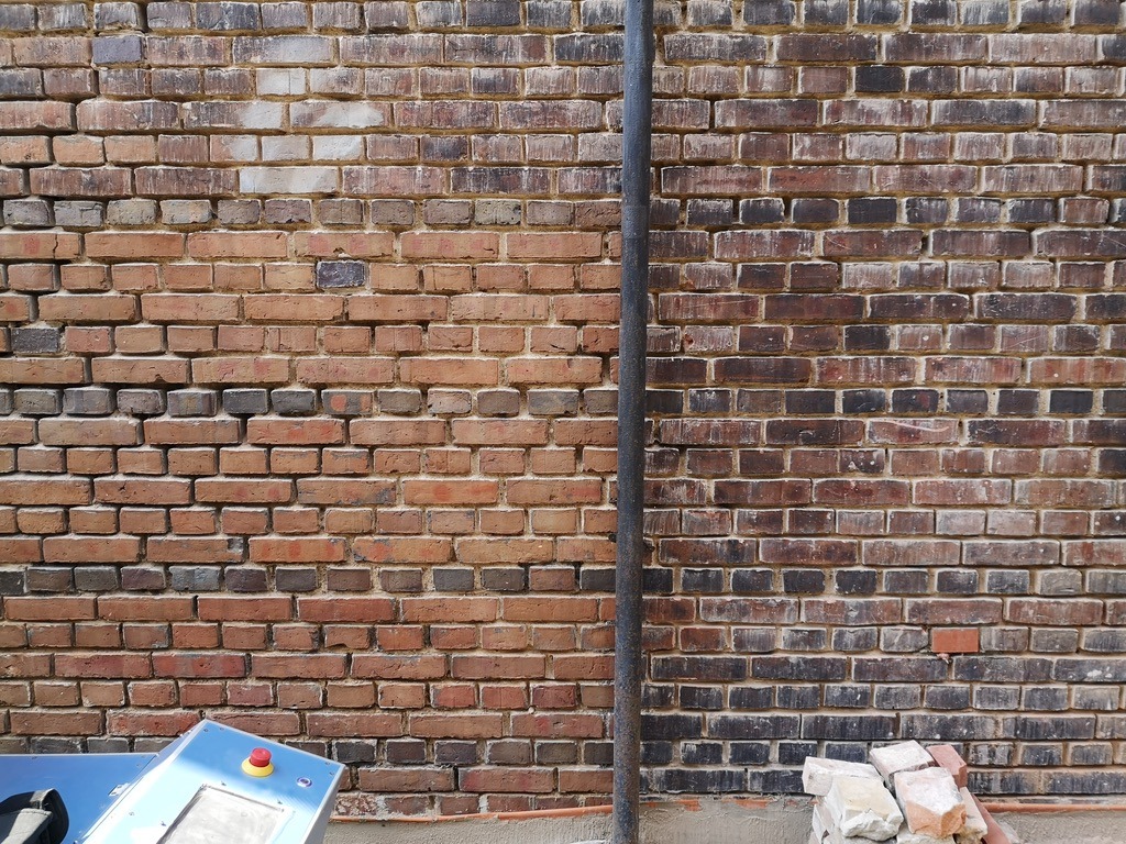 Mieszana ściana z czerwonej cegły z pionową rurą, fragmentem niebieskiego urządzenia z czerwonym przyciskiem i stertą cegieł u podstawy.