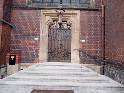 Wejście do budynku z czerwonej cegły, z kamiennymi schodami i ozdobnymi metalowymi drzwiami z lampionem powyżej.