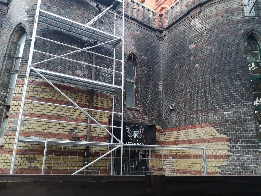 Fragment elewacji zabytkowego budynku z cegły, z widocznymi śladami zabrudzeń i osadzonym brudem, częściowo zakryty rusztowaniem i czarnymi drzwiami z białym logo.