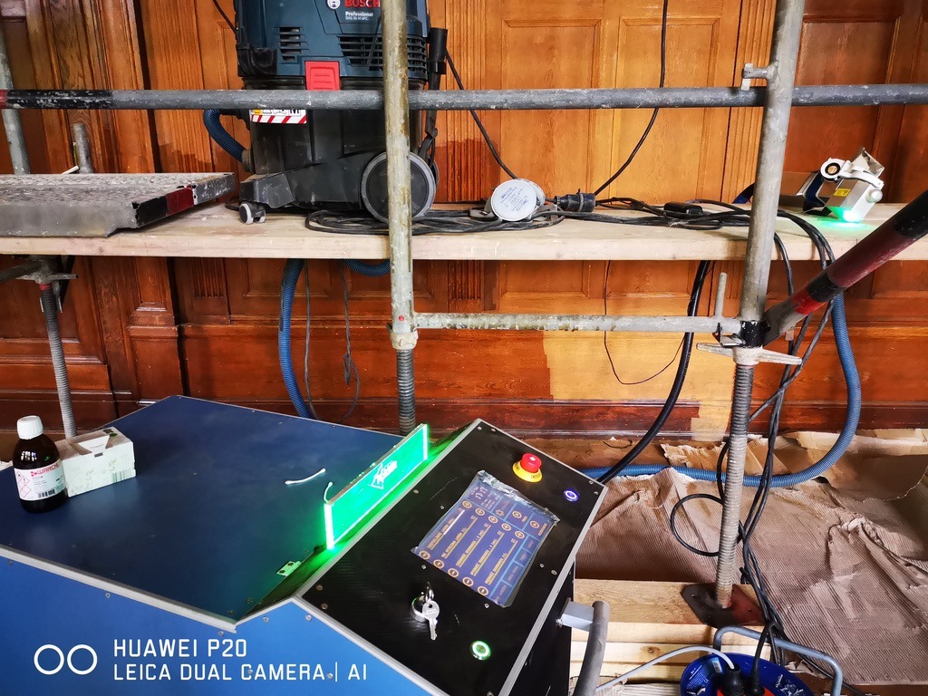 Fragment drewnianej boazerii w trakcie renowacji, widoczne rusztowanie, odkurzacz przemysłowy Bosch, urządzenie sterujące z kluczykiem i butelka z preparatem, fragment oczyszczonej powierzchni.