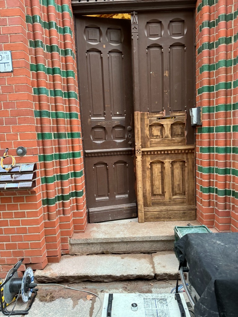 Otwarta, ciemnobrązowa, zabytkowa brama wejściowa z częściowo odnowionym fragmentem drzwi, ukazującym jaśniejszy odcień drewna, na tle ceglanej fasady z zielonymi zdobieniami.