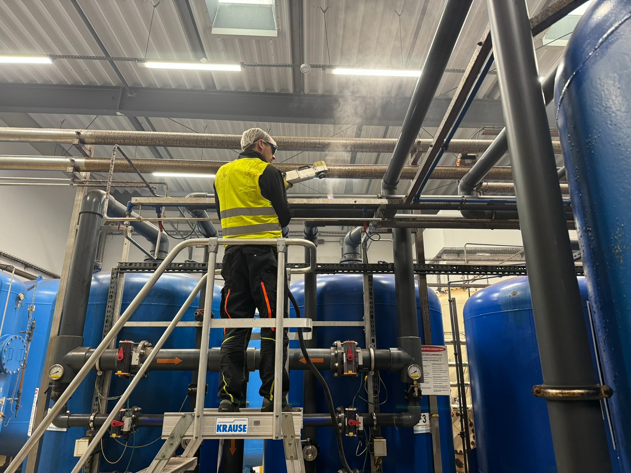Pracownik w odzieży ochronnej, stojący na aluminiowej drabinie Krause, czyści parą rury w instalacji przemysłowej z niebieskimi zbiornikami.