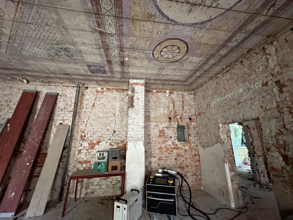 Wnętrze pomieszczenia w trakcie renowacji, widoczne odsłonięte ceglane ściany, fragmenty dekoracyjnego sufitu z malowidłami, sprzęt do prac konserwatorskich oraz stare skrzynki na stole.