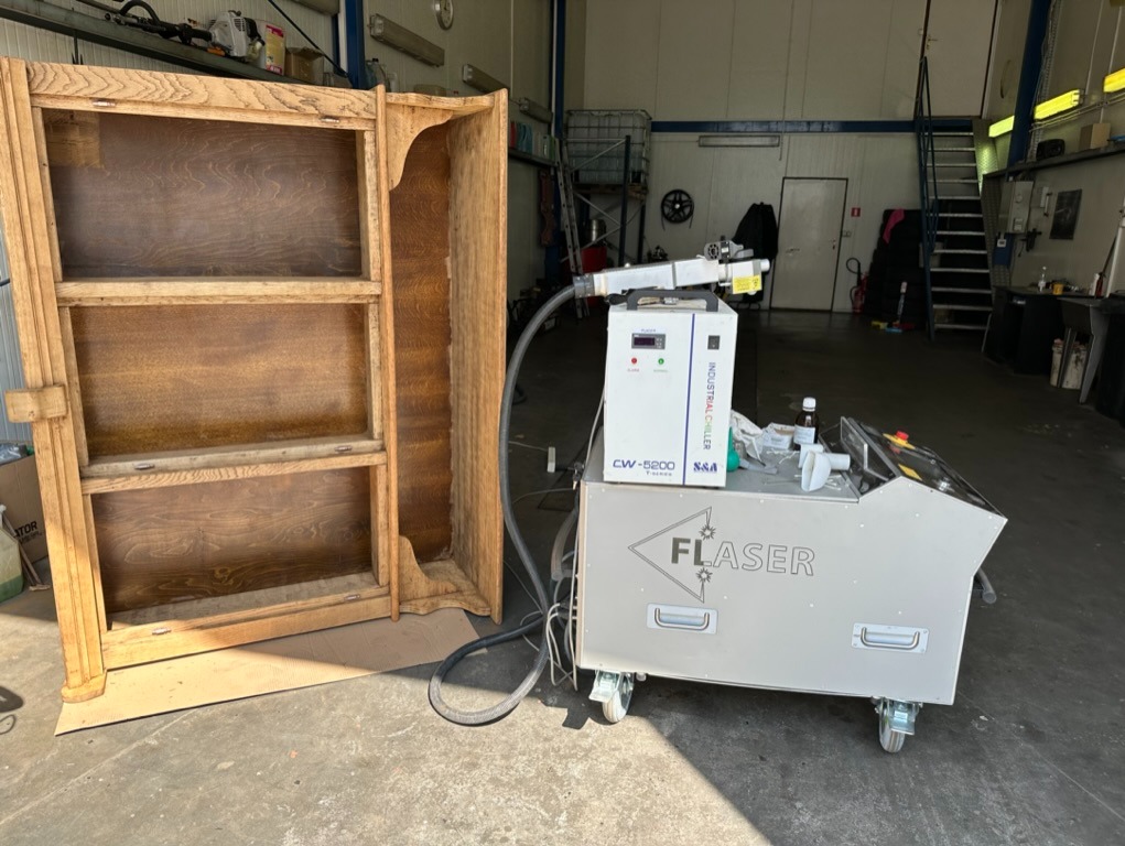 Drewniana szafka bez drzwi, z trzema półkami, obok maszyna przemysłowa z napisem FLaser, prawdopodobnie do czyszczenia lub renowacji drewna.