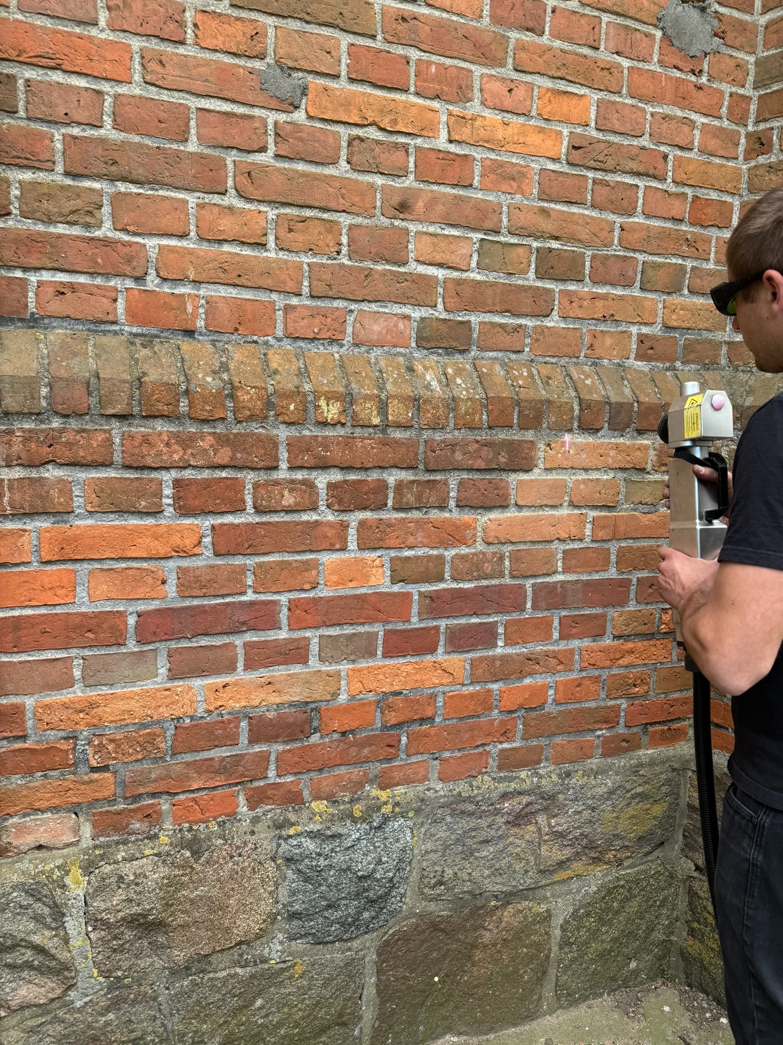 Pracownik w okularach ochronnych skanuje czerwoną ceglaną ścianę z kamienną podstawą przy użyciu urządzenia laserowego emitującego czerwoną wiązkę.