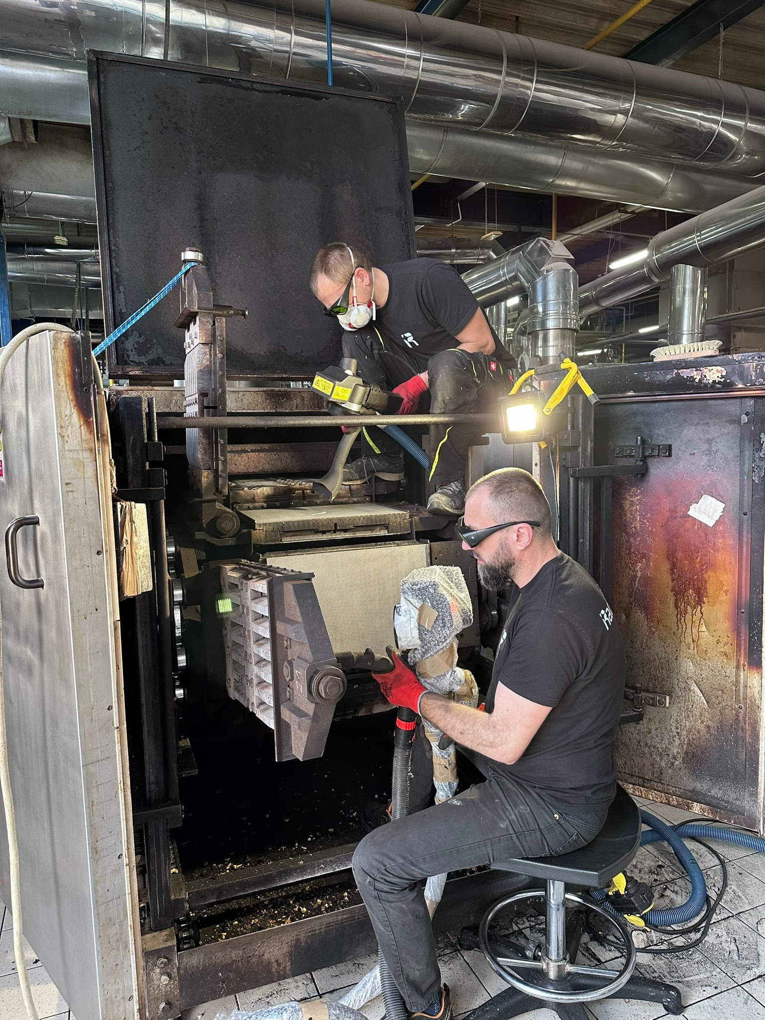 Dwóch mężczyzn w okularach ochronnych i czerwonych rękawicach czyści przemysłowo wnętrze dużej, metalowej maszyny, prawdopodobnie pieca przemysłowego, przy użyciu odkurzacza i latarki.