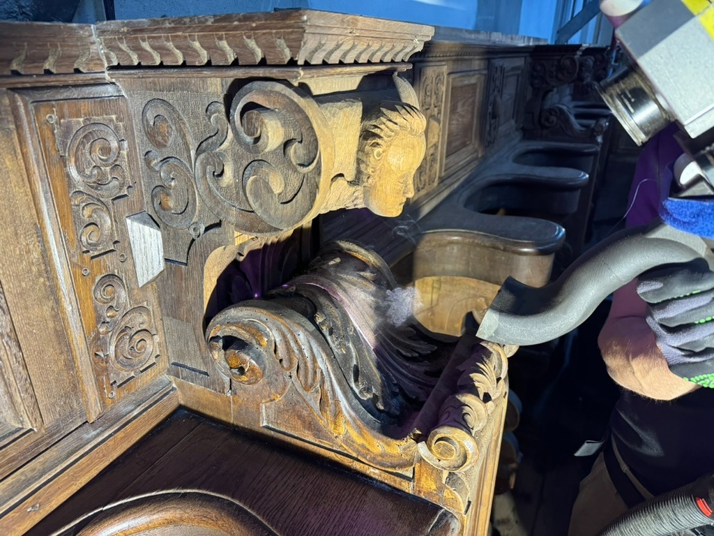 Szczegółowe czyszczenie rzeźbionej, drewnianej ławy kościelnej z użyciem odkurzacza przemysłowego, widoczne drobinki kurzu unoszące się w powietrzu.