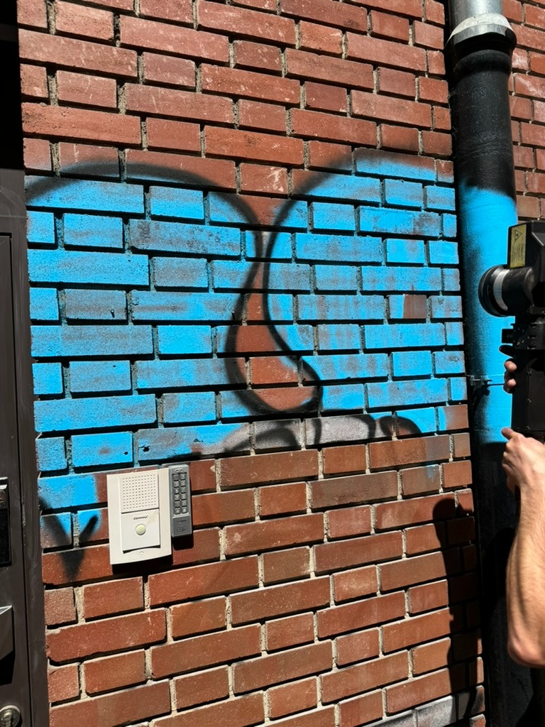 Ceglana ściana z pozostałościami niebieskiej farby i czarnym graffiti, obok rury spustowej i domofonu, w trakcie czyszczenia elewacji, z widocznym fragmentem urządzenia czyszczącego w prawym dolnym...