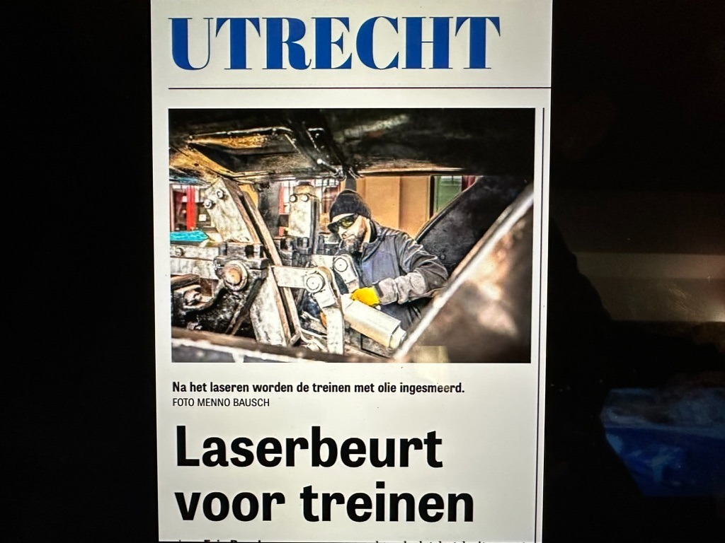 Zdjęcie artykułu prasowego z nagłówkiem 'UTRECHT' przedstawiające mężczyznę w okularach ochronnych i rękawicach, pracującego nad metalową konstrukcją przypominającą podwozie pociągu. W tle widoczne...